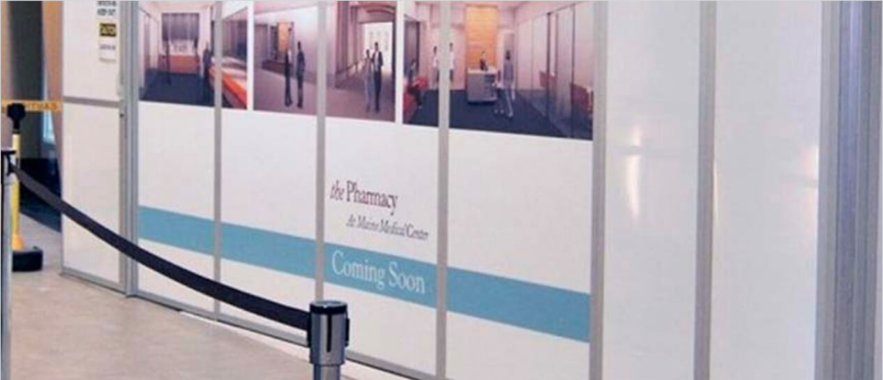 Temporary construction barrier with 'Coming Soon' signage for a new pharmacy at a medical center, featuring interior concept renderings and crowd control stanchions