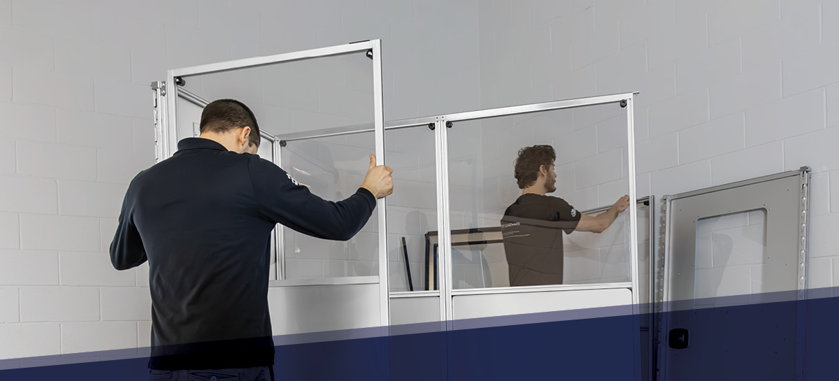 Two technicians assembling a modular containment wall system with transparent panels in a commercial facility. Designed for quick setup in construction zones, healthcare renovations, and cleanroom environments to control dust and airborne contaminants.