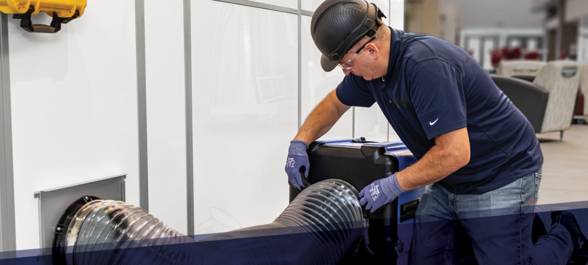 Technician setting up a HEPA air filtration system using flexible ducting, connected to a temporary containment barrier in a healthcare or commercial facility setting.