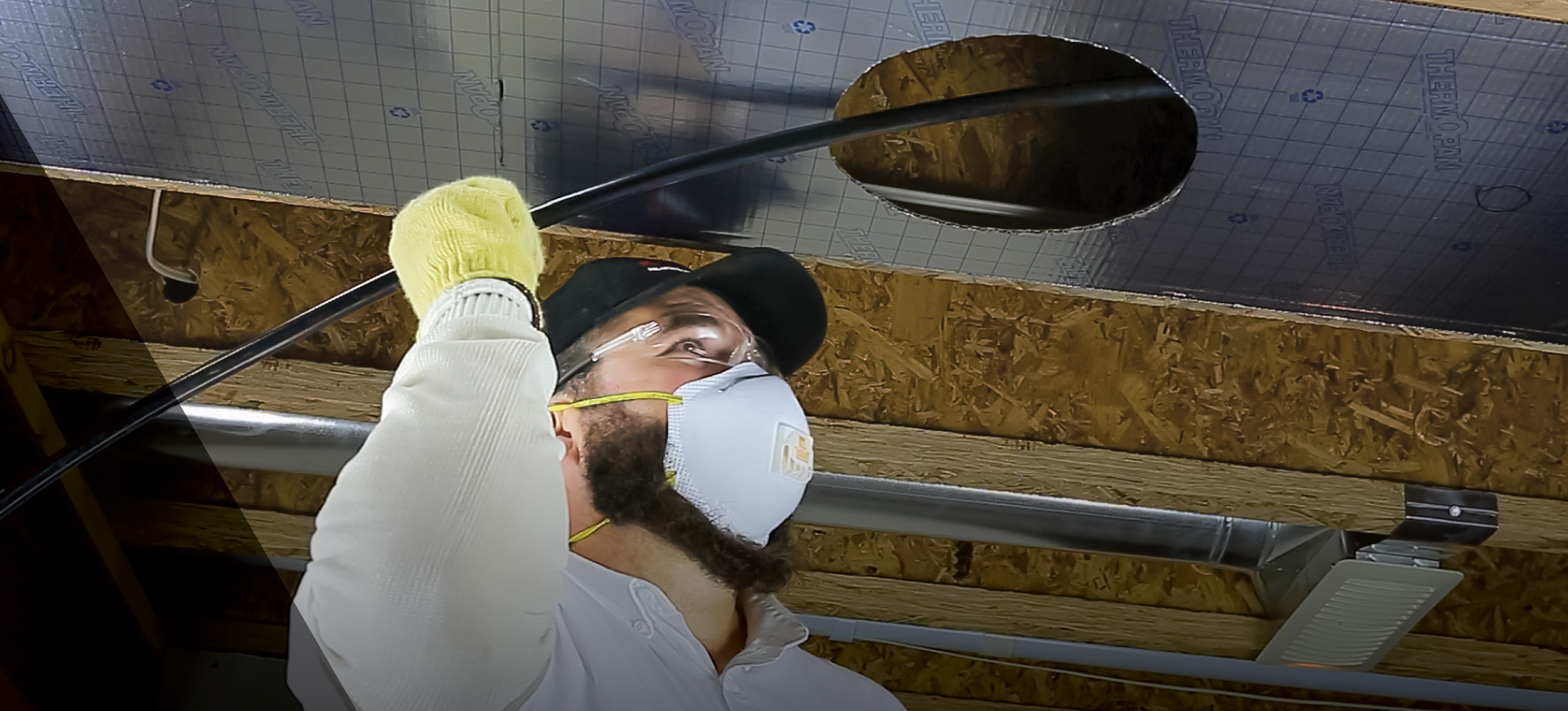 Un technicien portant un masque, des lunettes de protection et des gants utilise un fouet à air comprimé pour nettoyer l'intérieur d'un conduit d'air dans un plafond suspendu, dans le cadre du processus de nettoyage de conduits DUCT-PRO®.